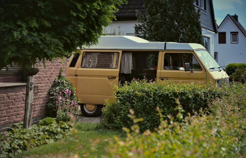 A 1980s VW bus, yellow below with a white high roof, parked behind a hedge